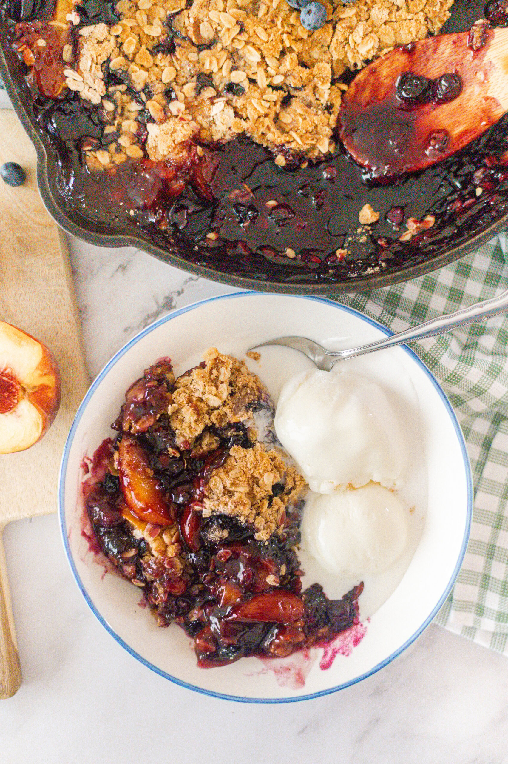 A bowl of Cast Iron Skillet Peach Blueberry Crisp is served with two scoops of vanilla ice cream, beside the skillet of crisp and a sliced peach on a cutting board. A green checkered towel is partially visible.