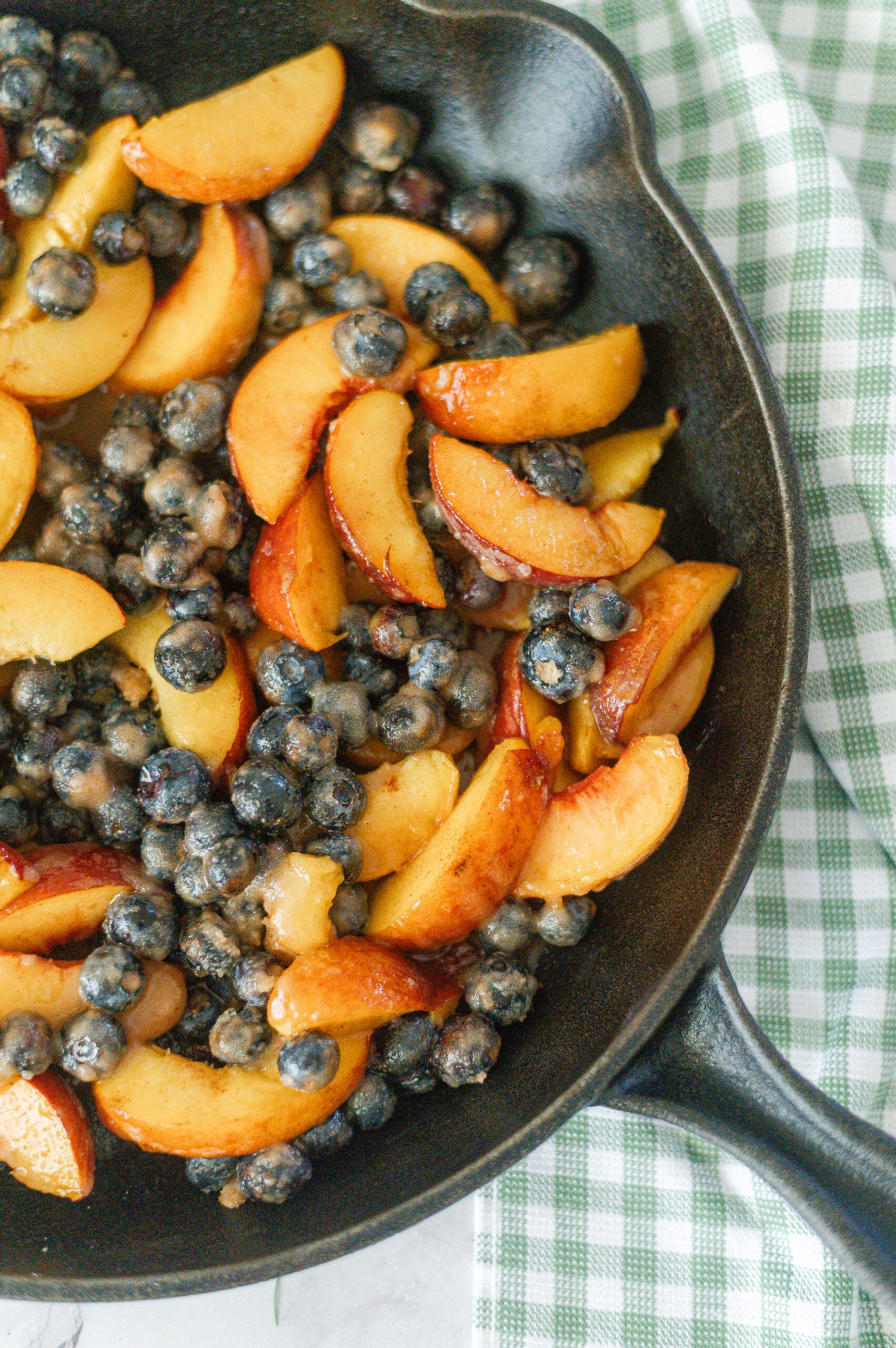 A Cast Iron Skillet Peach Blueberry Crisp, partially cooked with sliced peaches and blueberries, rests on a green and white checkered towel.