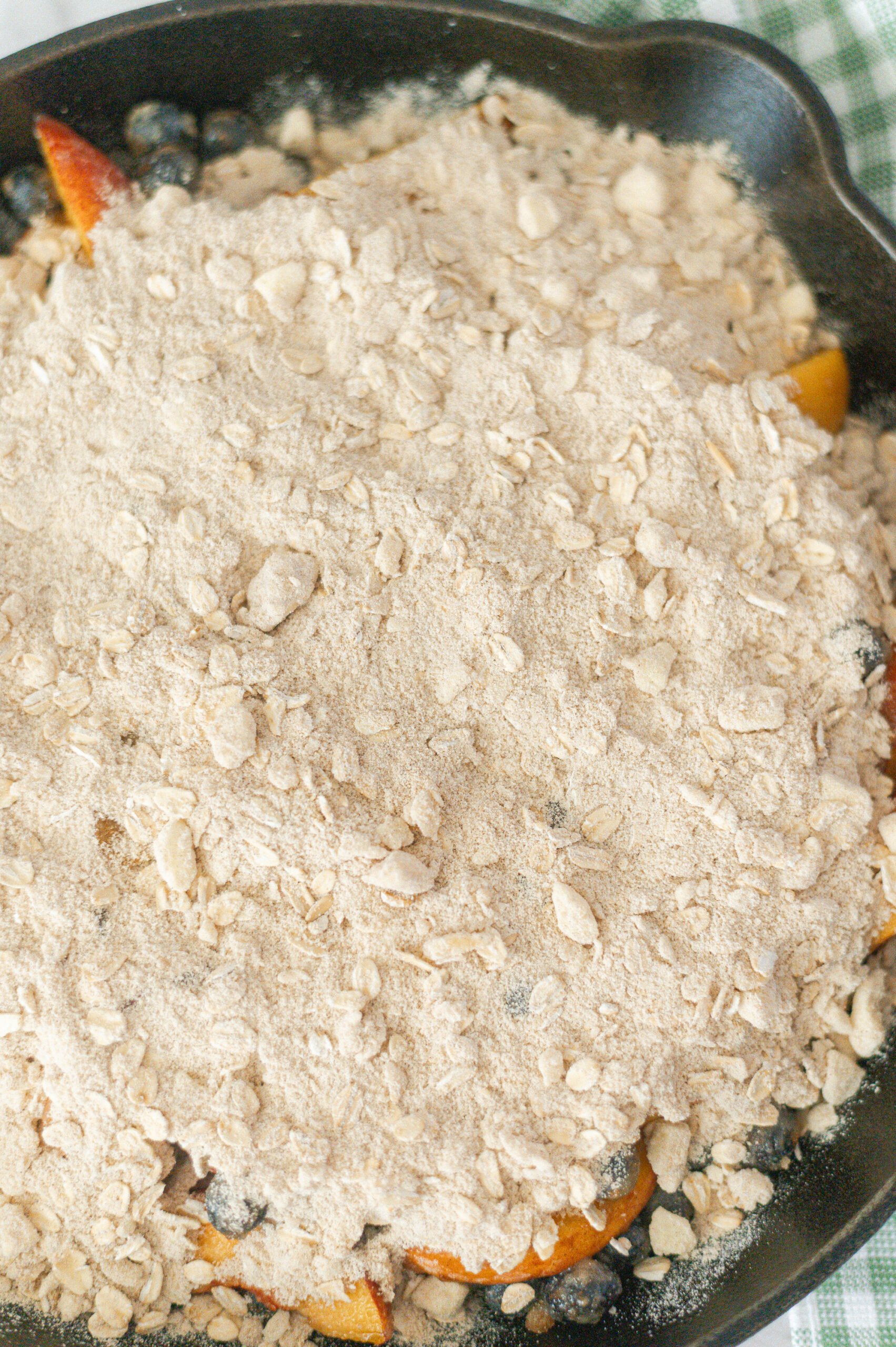 A close-up of an unbaked Cast Iron Skillet Peach Blueberry Crisp, topped with a layer of oat and flour crumble, with juicy blueberries and peach slices peeking through underneath.
