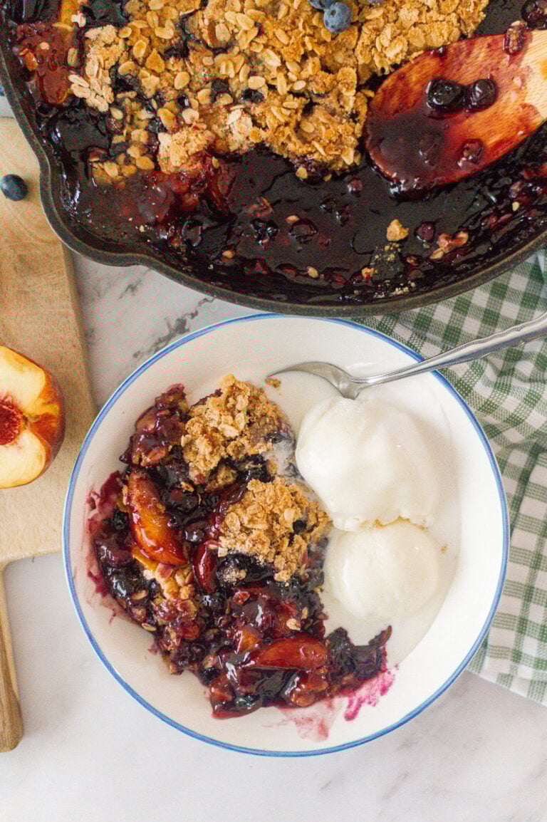 A bowl with a serving of Cast Iron Skillet Peach Blueberry Crisp and two scoops of vanilla ice cream, next to a skillet filled with more crisp, fresh peaches, and a green checkered napkin.