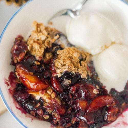 A bowl of Cast Iron Skillet Peach Blueberry Crisp with a golden oat topping, served with two scoops of vanilla ice cream. A spoon rests in the bowl, and the dessert has a mix of berry and stone fruit filling.
