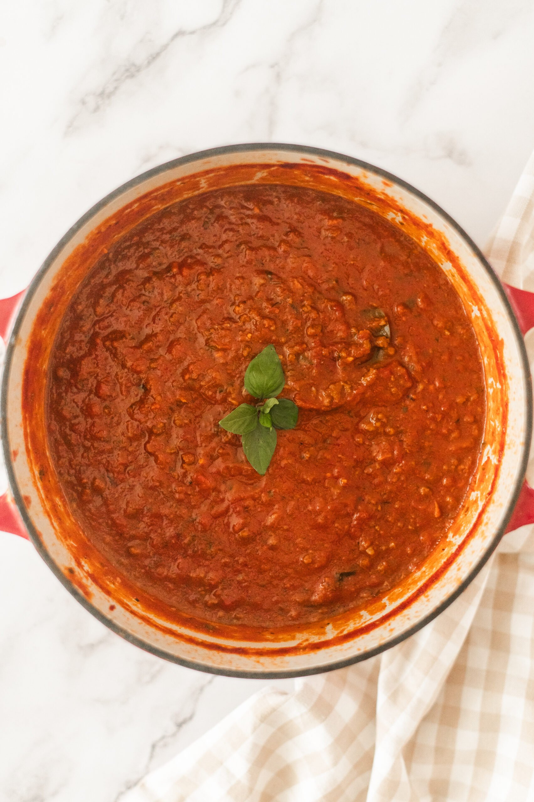 A pot of thick, red pasta sauce garnished with fresh basil leaves sits on a marble countertop next to a beige checked kitchen towel.