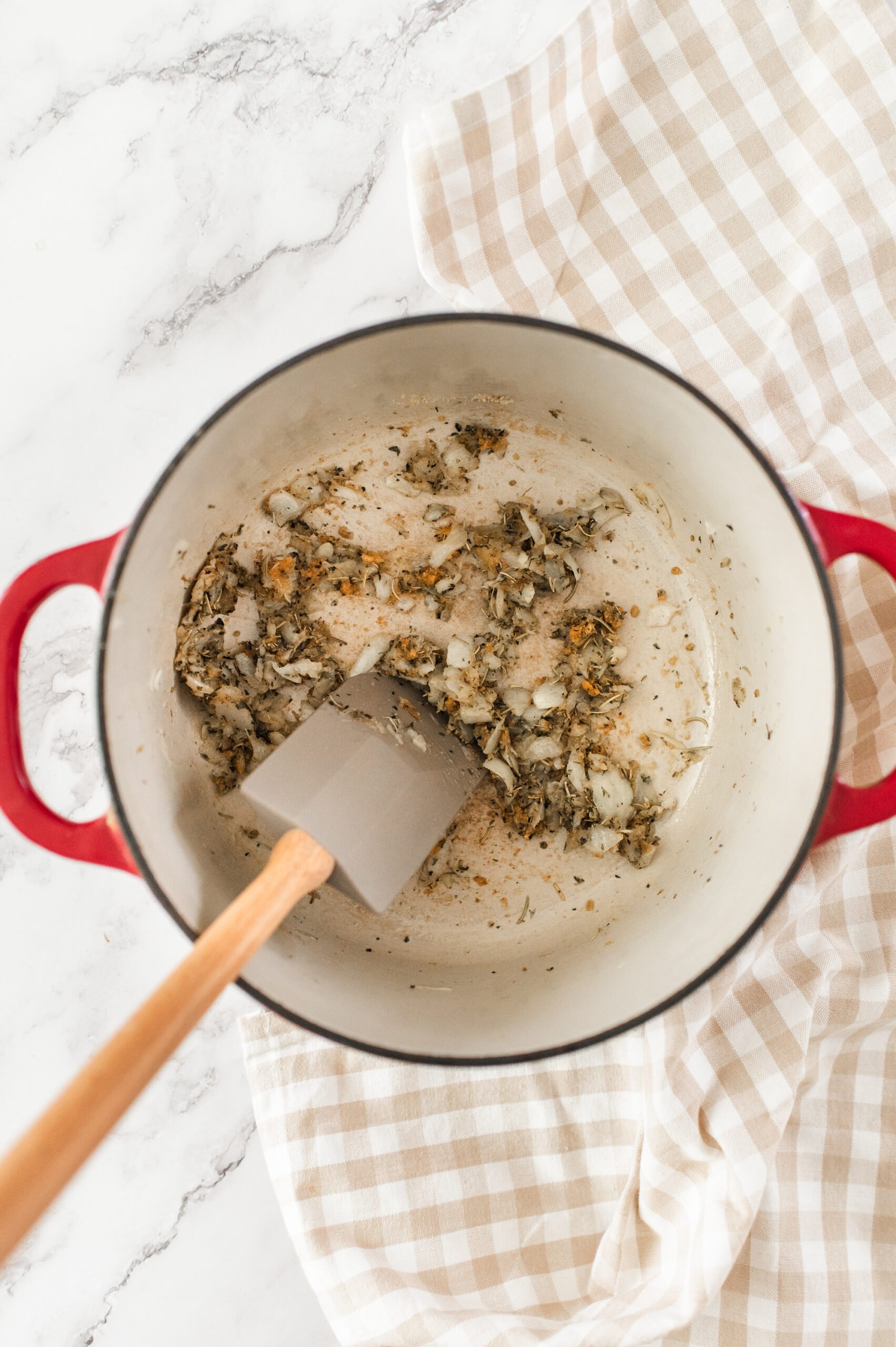 A red Dutch oven with sautéed onions and herbs being stirred with a spatula, placed on a light checkered cloth on a marble surface.