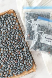 A baking sheet covered with a single layer of blueberries sits next to two labeled freezer bags filled with blueberries. A white kitchen towel is in the background on a marble surface.