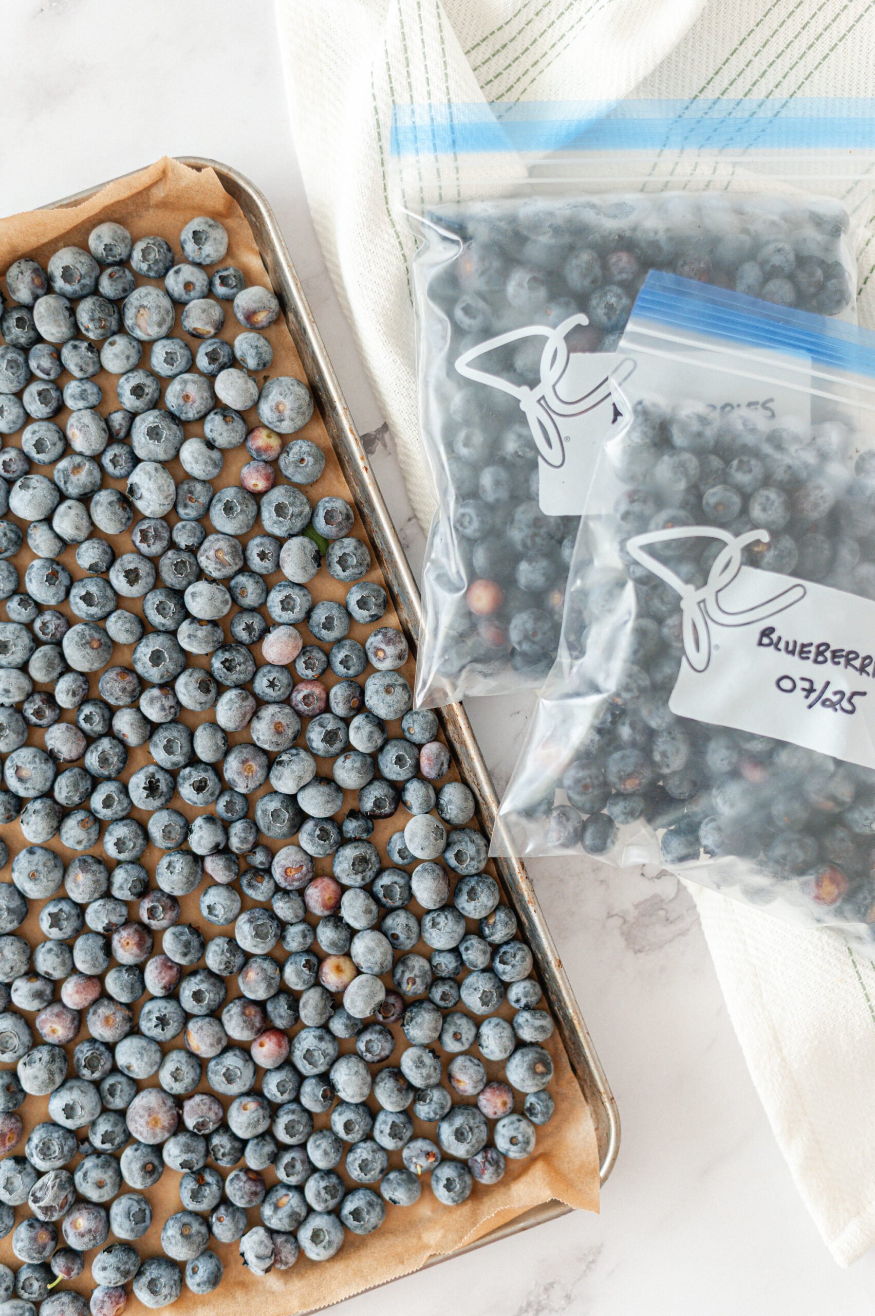 A baking sheet covered with a single layer of blueberries sits next to two labeled freezer bags filled with blueberries. A white kitchen towel is in the background on a marble surface.