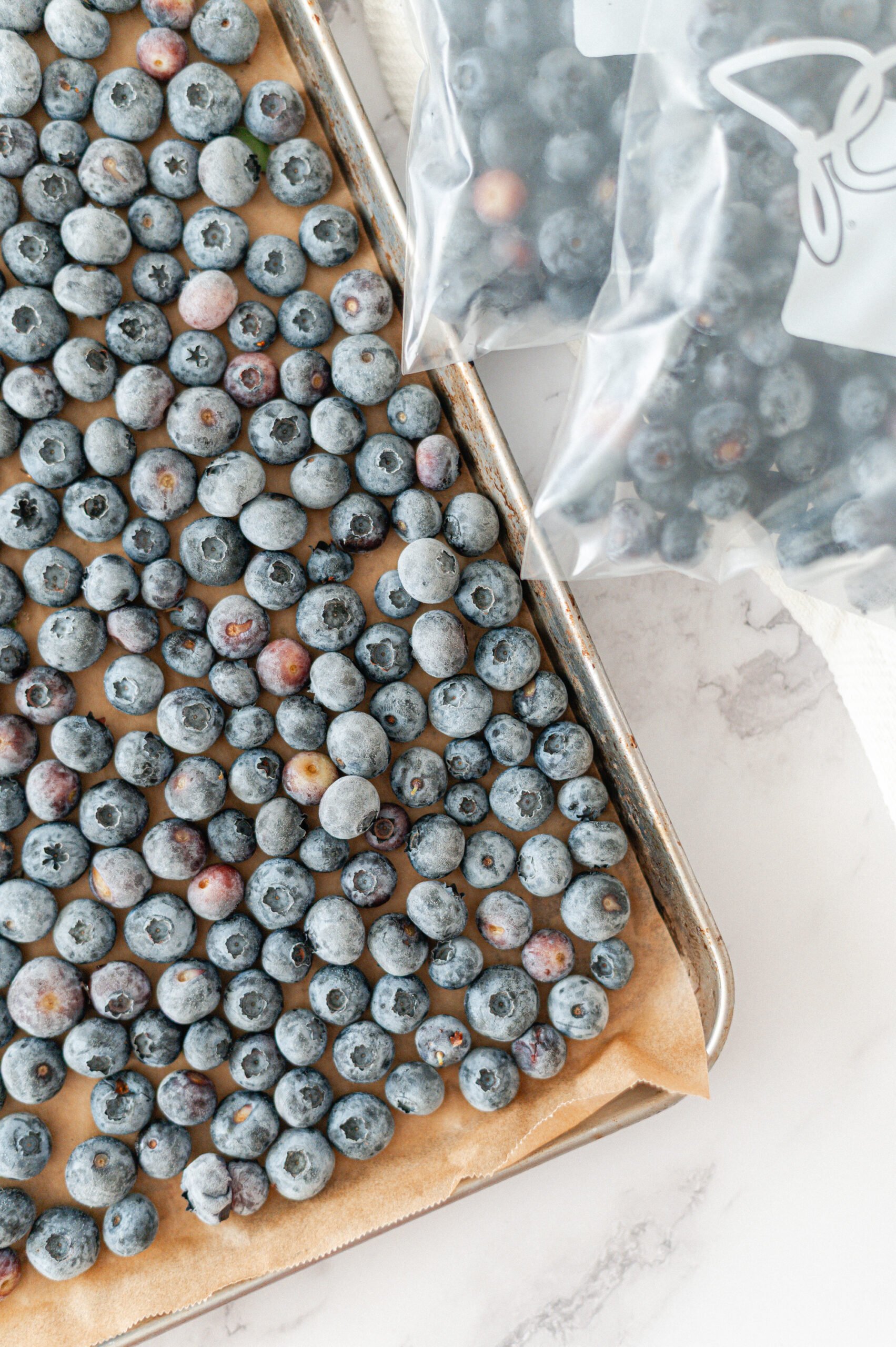A baking sheet lined with parchment paper is covered with frozen blueberries. In the background, two plastic bags filled with more blueberries are partially visible on a marble surface.