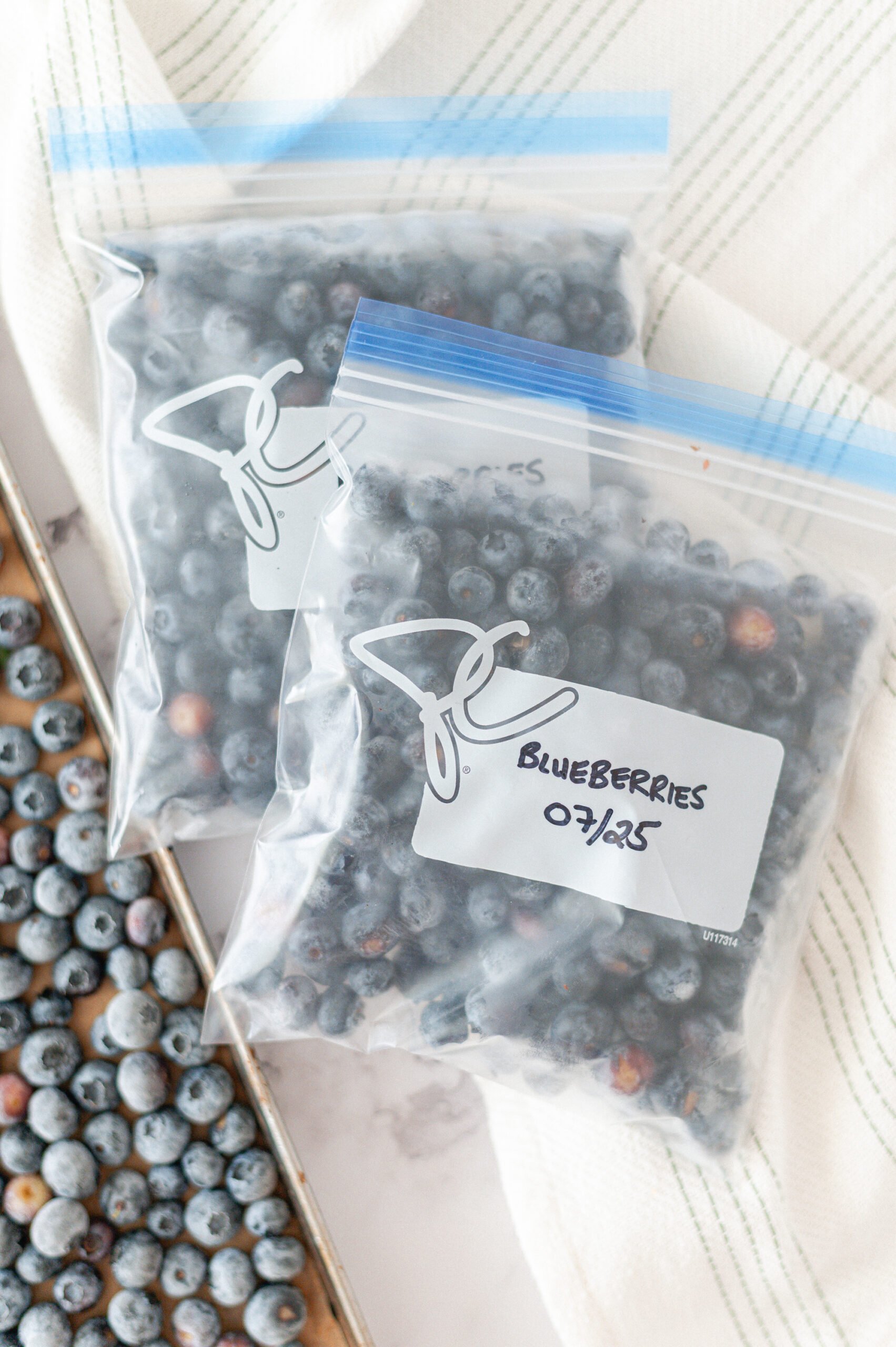 Two labeled freezer bags filled with blueberries are on a white towel. A baking sheet with loose blueberries is next to the bags. The bags are labeled Blueberries 07/25 with a marker.