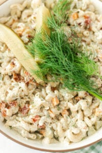 A bowl of creamy pasta salad with chopped vegetables, topped with two pickle spears and a sprig of fresh dill.