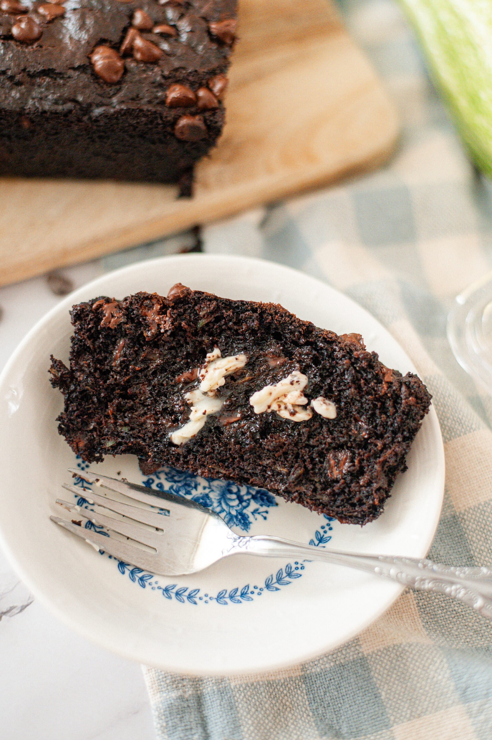 A slice of healthy chocolate zucchini bread topped with a pat of butter on a decorative plate with a fork beside it, showcasing its rich, fudgy texture.