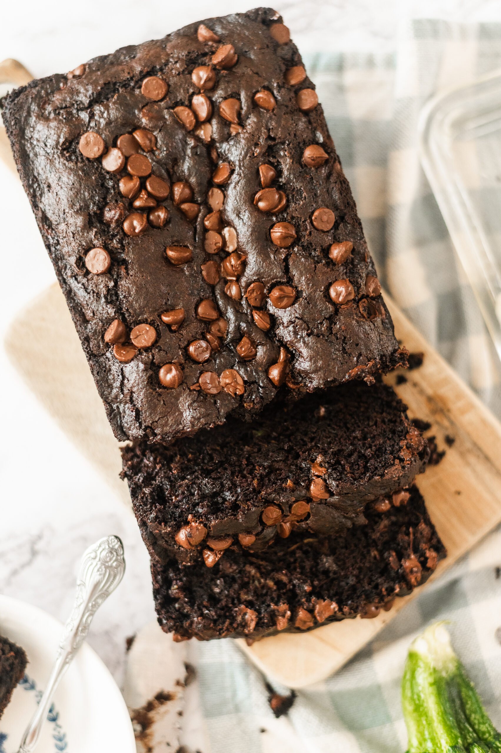 A loaf of chocolate zucchini bread topped with chocolate chips, sliced on a wooden cutting board, with a zucchini in the background.