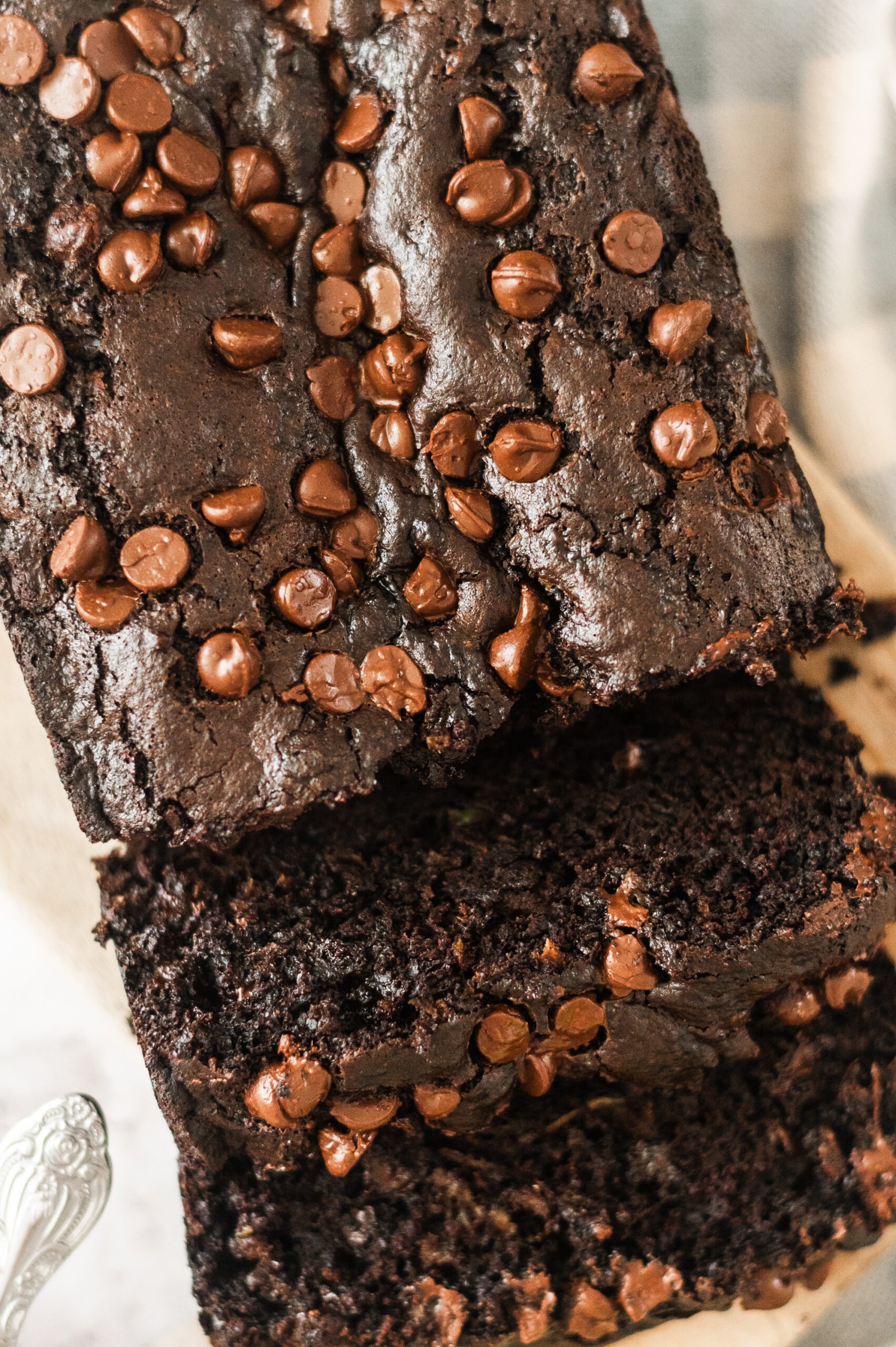 Close-up of a moist chocolate zucchini bread topped with chocolate chips, displaying a rich, fudgy texture. The loaf is sliced to reveal its dark, soft interior, ideal for a dessert or snack.