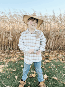 A smiling child dressed as a scarecrow stands on grass in front of a cornfield, wearing a straw hat, plaid shirt, jeans, boots, and face paint with a drawn-on smile and stitches.