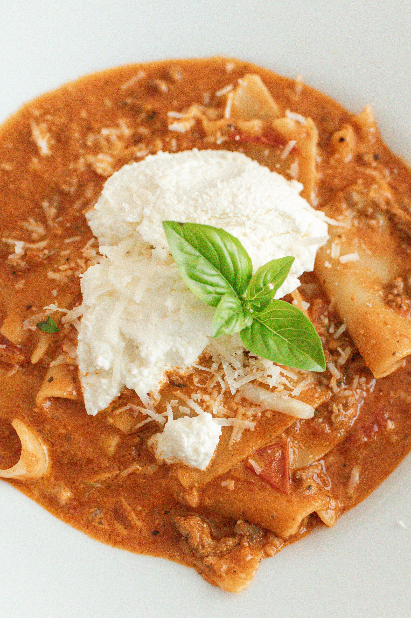 A close-up of creamy lasagna soup topped with a dollop of ricotta cheese, grated Parmesan, and a fresh basil sprig, served in a white bowl.