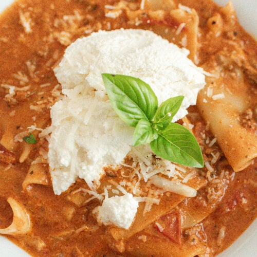A close-up of creamy lasagna soup topped with a dollop of ricotta cheese, grated parmesan, and a fresh basil leaf, served in a white bowl.