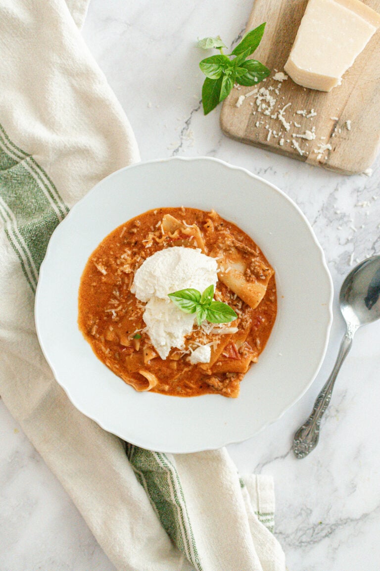 A bowl of lasagna soup topped with a dollop of ricotta cheese and fresh basil, set on a white surface with a striped towel, spoon, grated cheese, and a small basil sprig nearby.