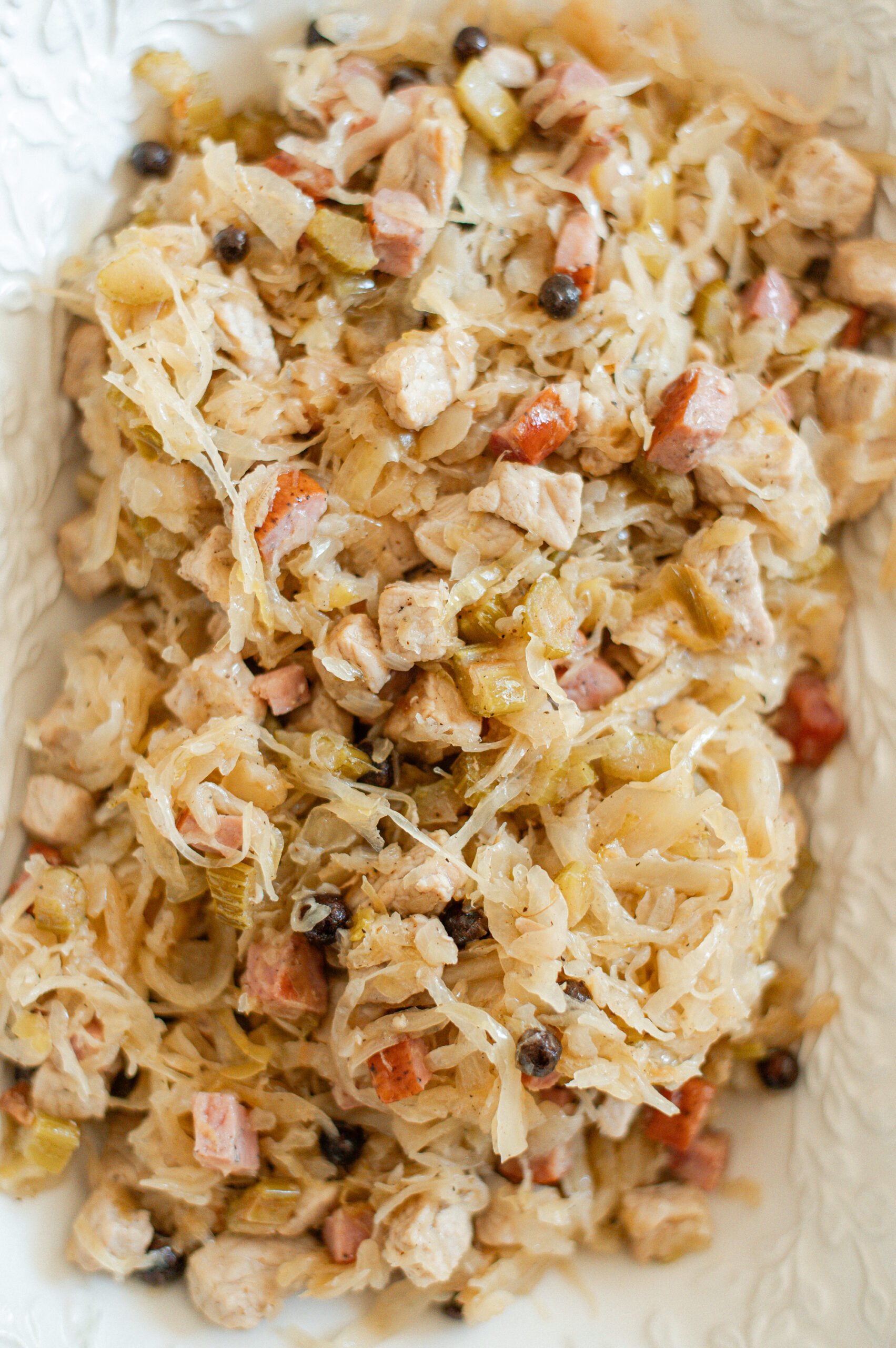 A close-up of a dish featuring shredded sauerkraut mixed with diced meats, sausage, and vegetables, seasoned with peppercorns and spices, served on a white plate.