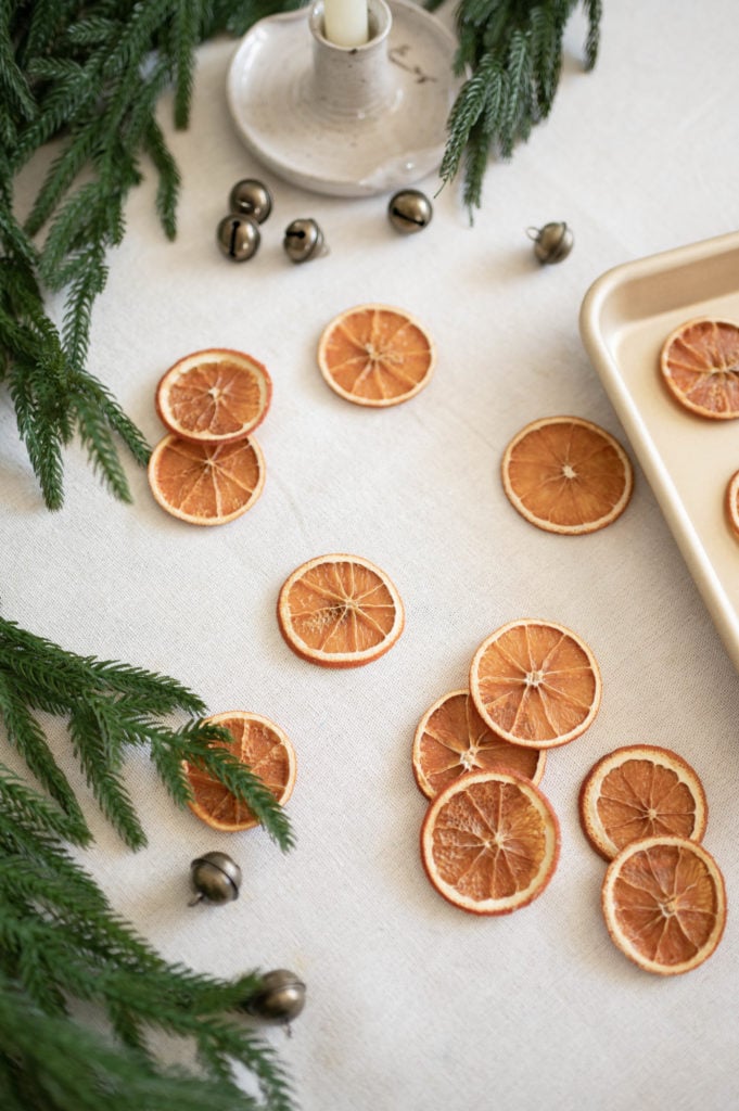 Dried orange slices are scattered on a white tablecloth, surrounded by green pine branches, small metallic bells, a beige tray with more orange slices, and a ceramic candle holder.