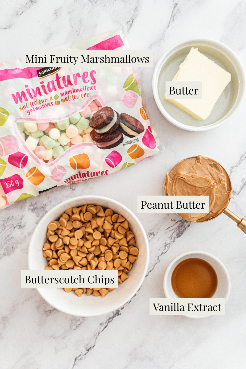 A marble surface displays mini fruity marshmallows, a dish of butter, a bowl of peanut butter, a bowl of butterscotch chips, and a small dish of vanilla extract, each labeled with text.