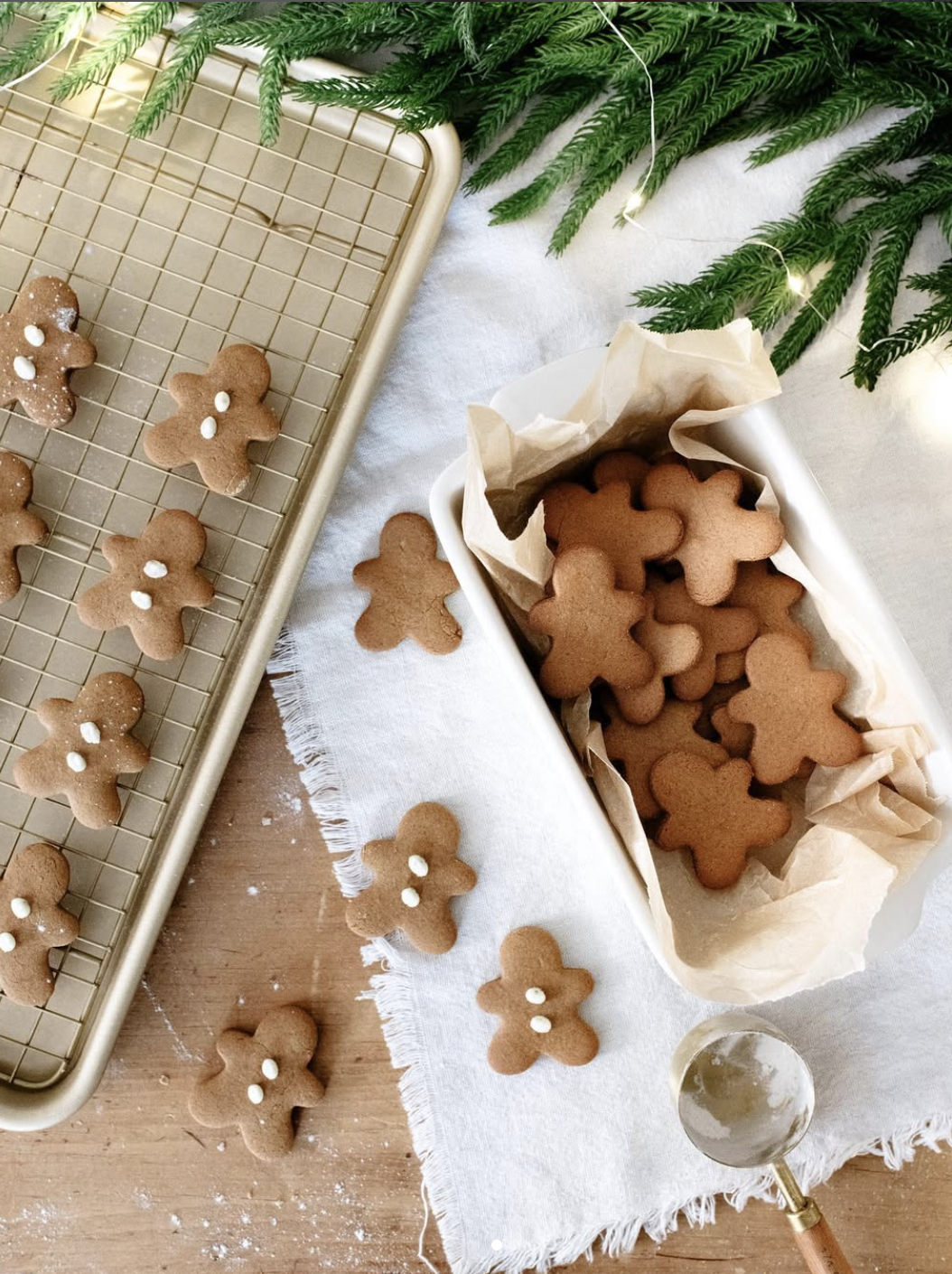 A tray and dish filled with gingerbread cookies shaped like people, some decorated with white icing, are on a linen cloth beside evergreen branches. A metal scoop and scattered flour are visible.