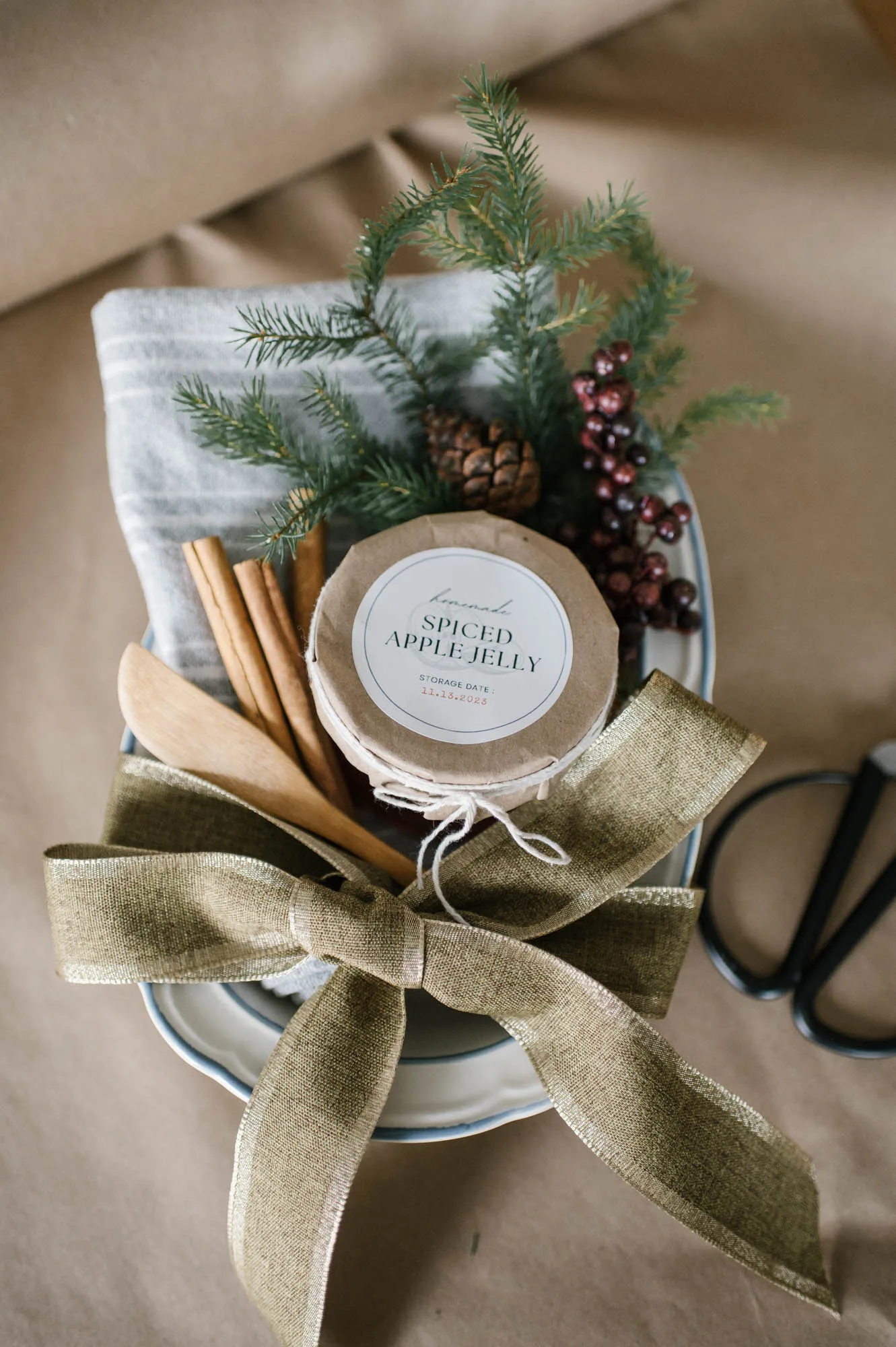 A gift basket with spiced apple jelly, a gray cloth, wooden utensils, cinnamon sticks, pine branches, berries, a pinecone, and a large burlap bow, all arranged on brown wrapping paper next to black scissors.