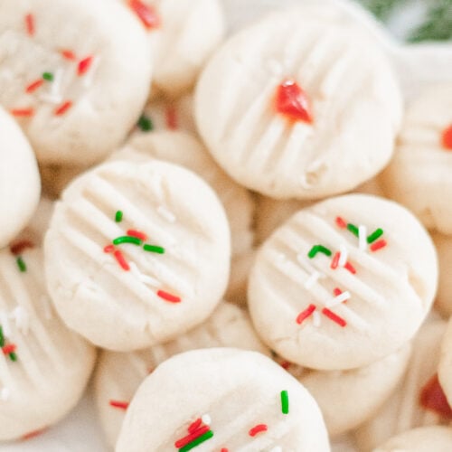 A plate of round, white Traditional Shortbread Cookies is topped with red and green sprinkles and red candied pieces, with some evergreen foliage in the background.