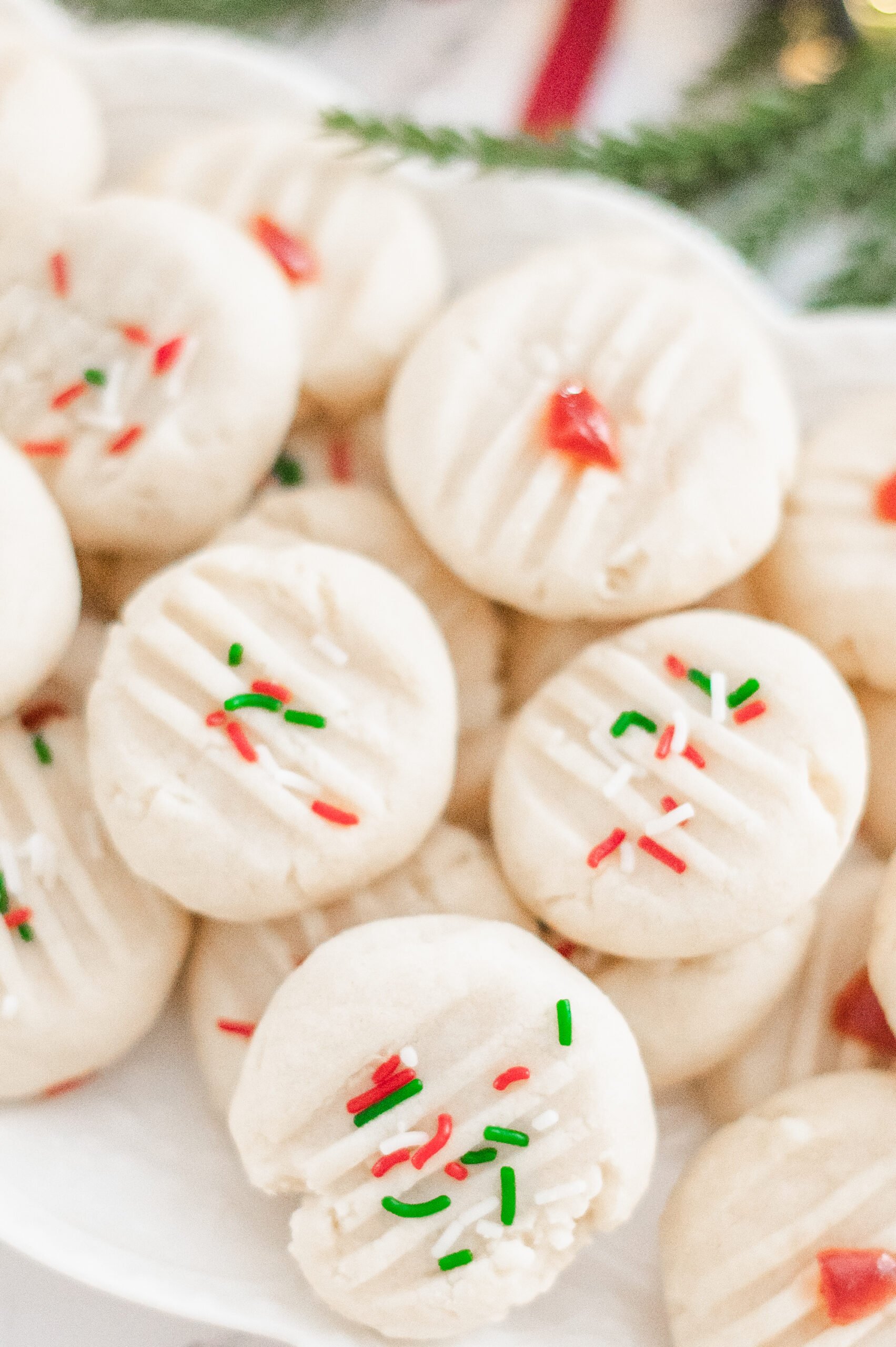 A plate of round, white Traditional Shortbread Cookies is topped with red and green sprinkles and red candied pieces, with some evergreen foliage in the background.