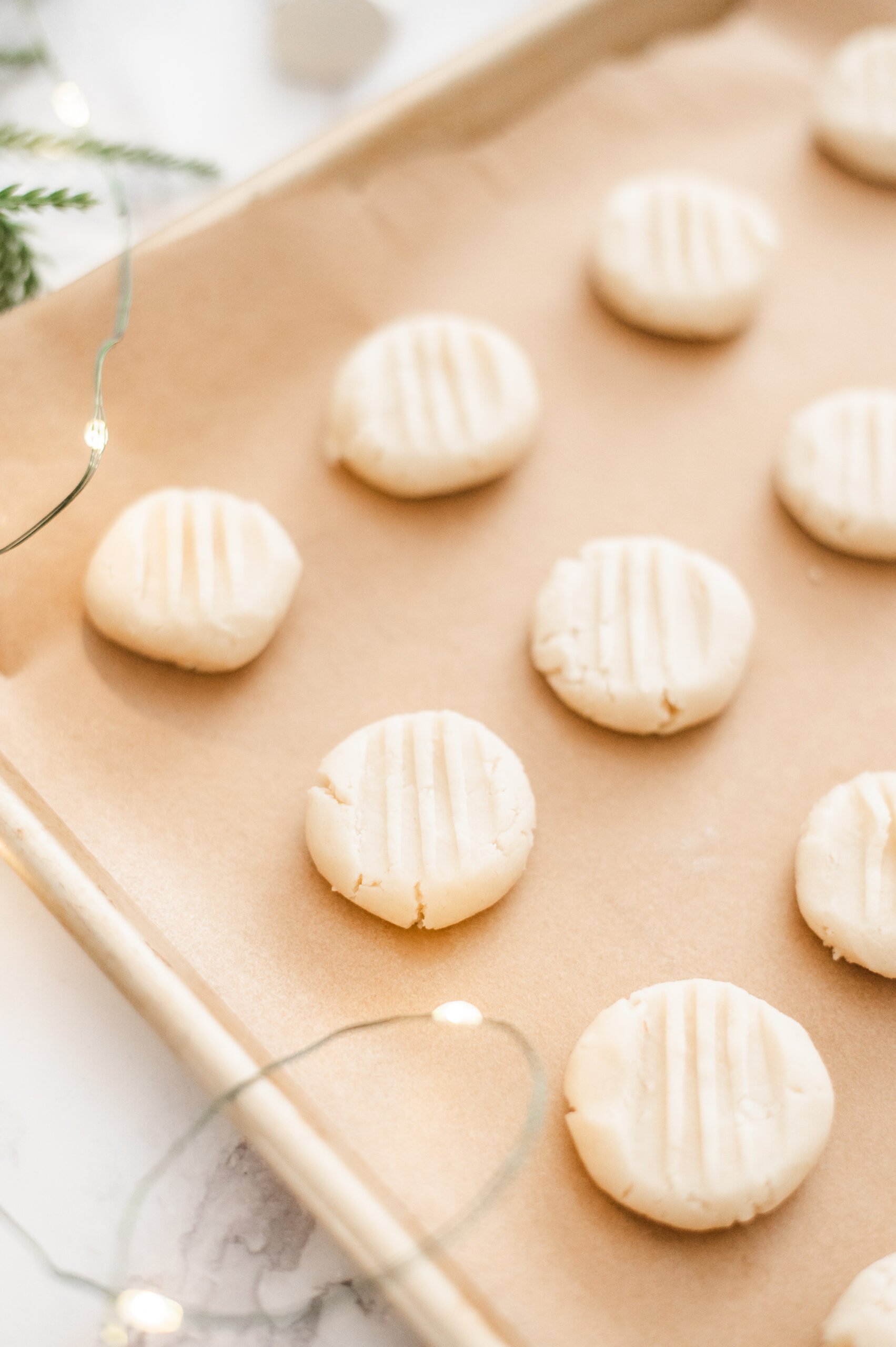Unbaked Traditional Shortbread Cookies with fork marks on top are arranged on a parchment-lined baking sheet. Holiday lights and greenery in the background create a festive atmosphere.