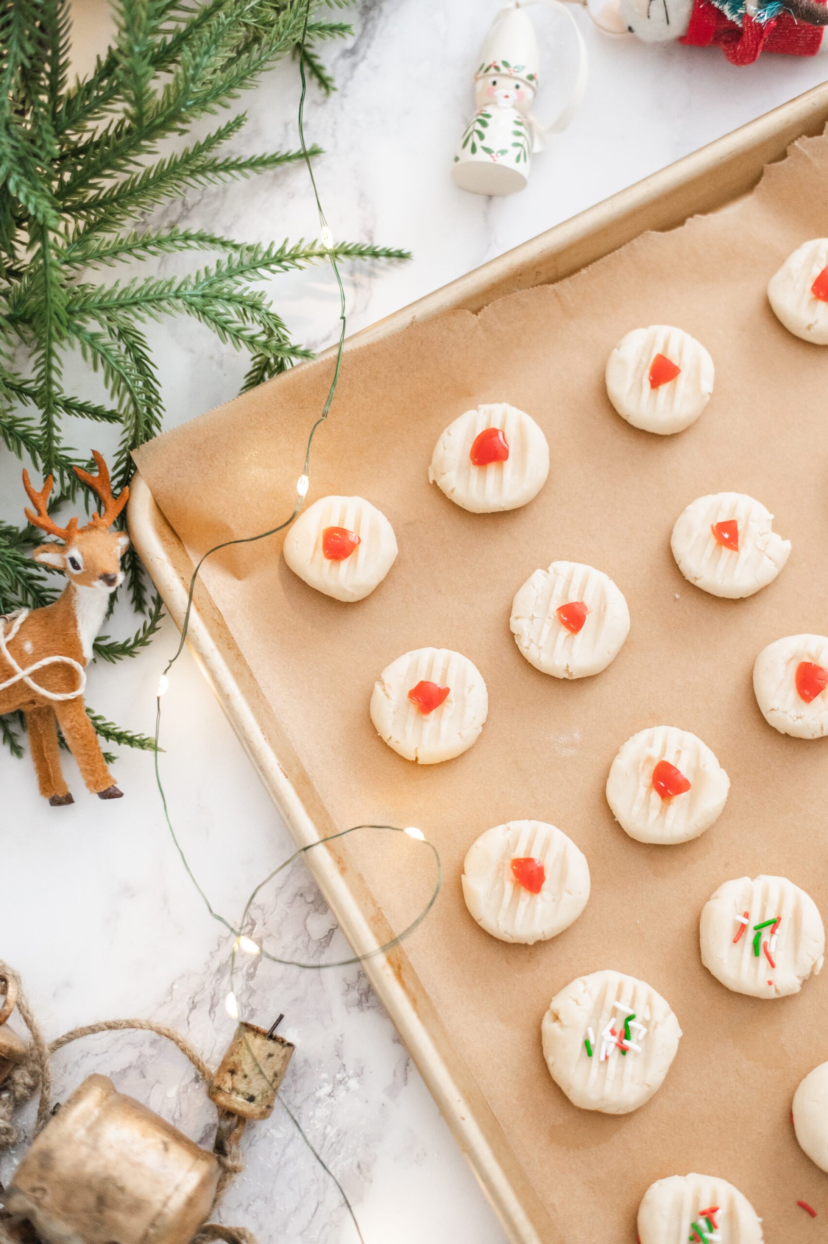 A baking tray lined with parchment paper holds unbaked Traditional Shortbread Cookies topped with red candied pieces. Nearby are Christmas decorations, pine branches, string lights, and a reindeer ornament on a marble surface.