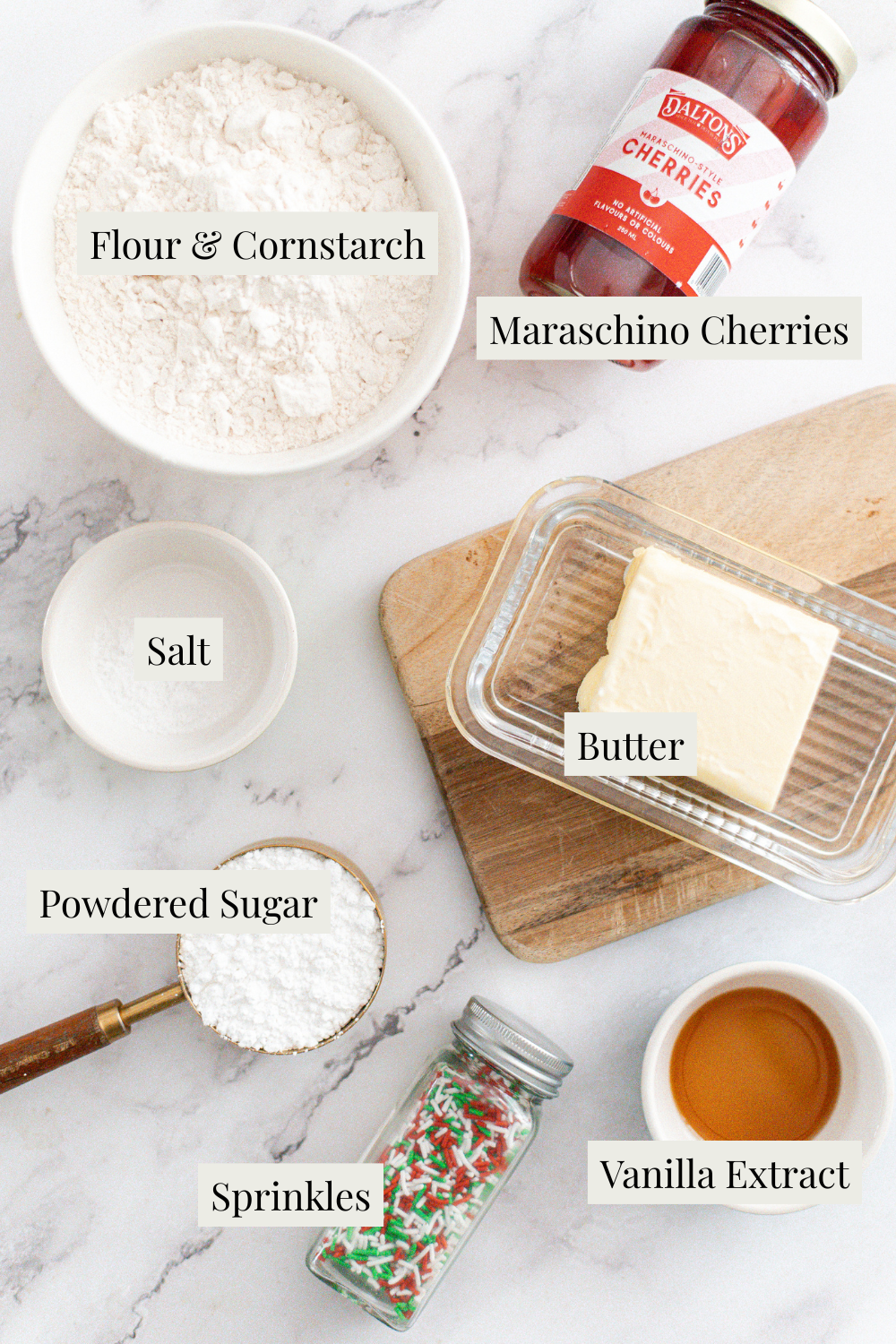 Top-down view of labeled baking ingredients for Traditional Shortbread Cookies on a white surface: flour & cornstarch, maraschino cherries, salt, butter, powdered sugar, sprinkles, and vanilla extract, some in bowls or jars.