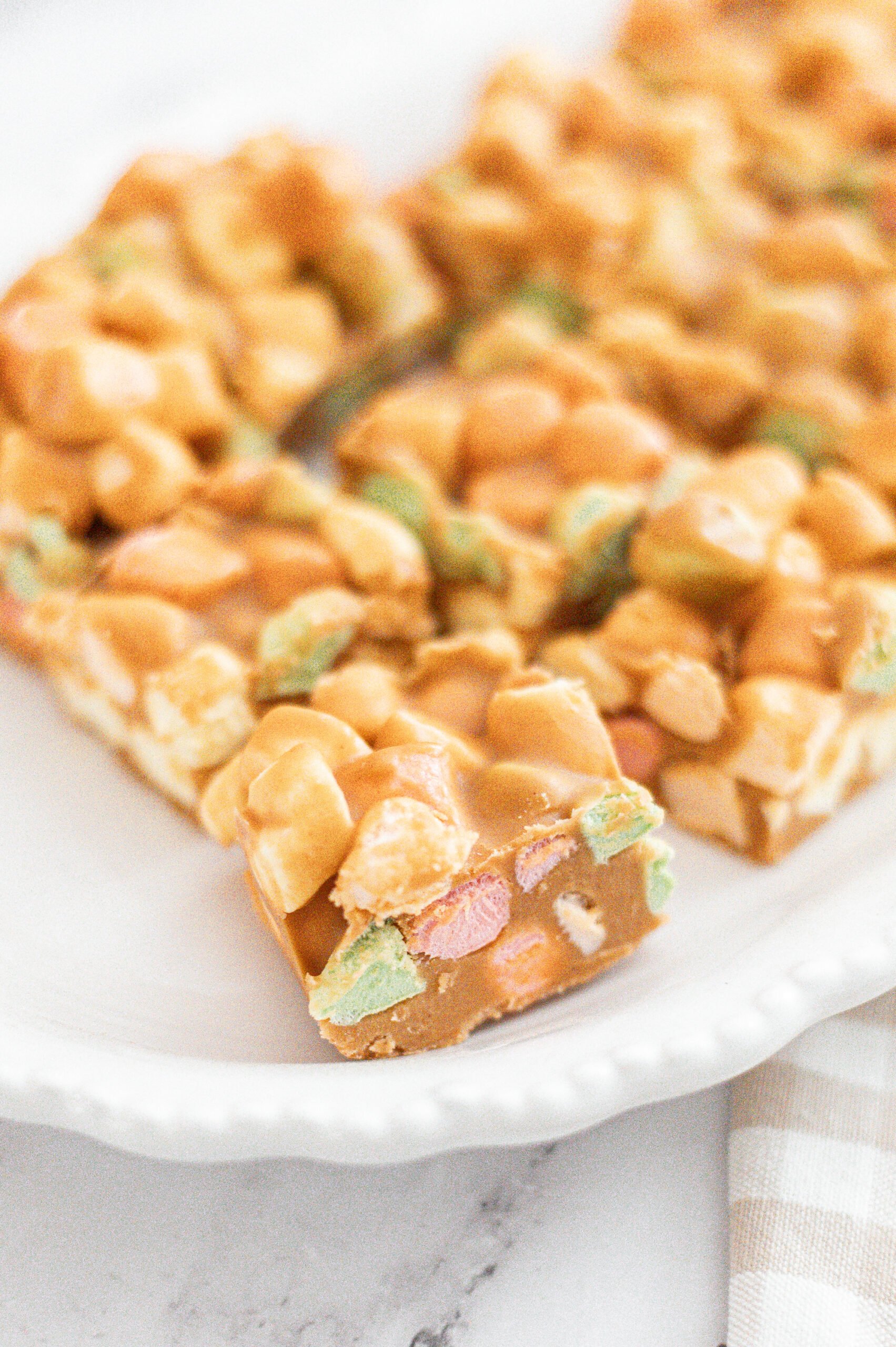 Close-up of a slice of butterscotch haystack bar made with colorful mini marshmallows and peanut butter, resting on a white plate. The bar has a smooth, glossy appearance and a chewy texture.
