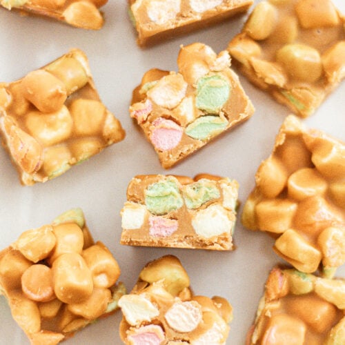 Squares of butterscotch marshmallow treats, showing colorful mini marshmallows and a golden, glossy coating, arranged on a white surface.