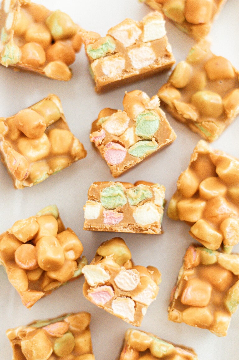 Squares of butterscotch marshmallow treats, showing colorful mini marshmallows and a golden, glossy coating, arranged on a white surface.