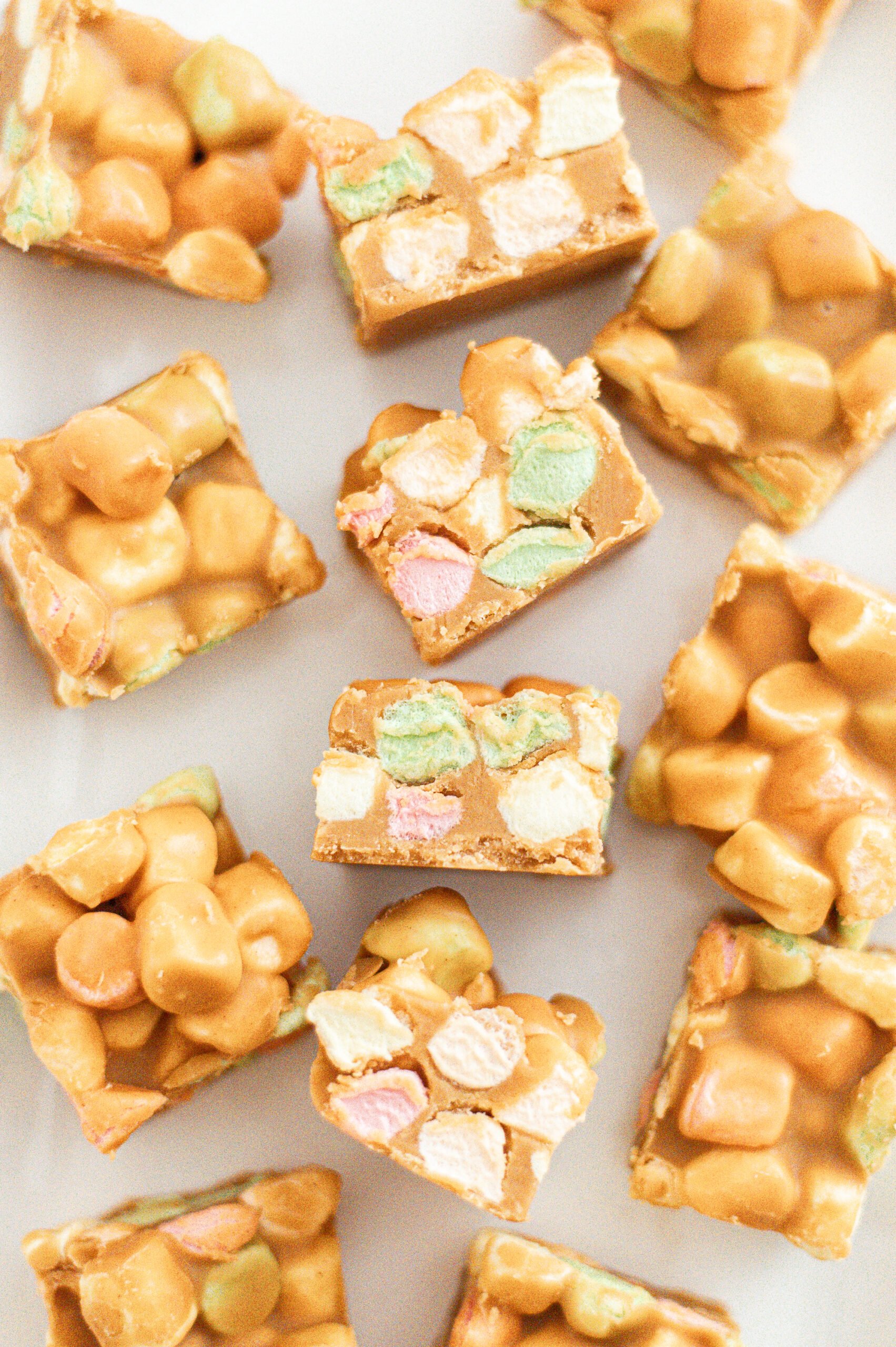 Squares of butterscotch marshmallow treats, showing colorful mini marshmallows and a golden, glossy coating, arranged on a white surface.