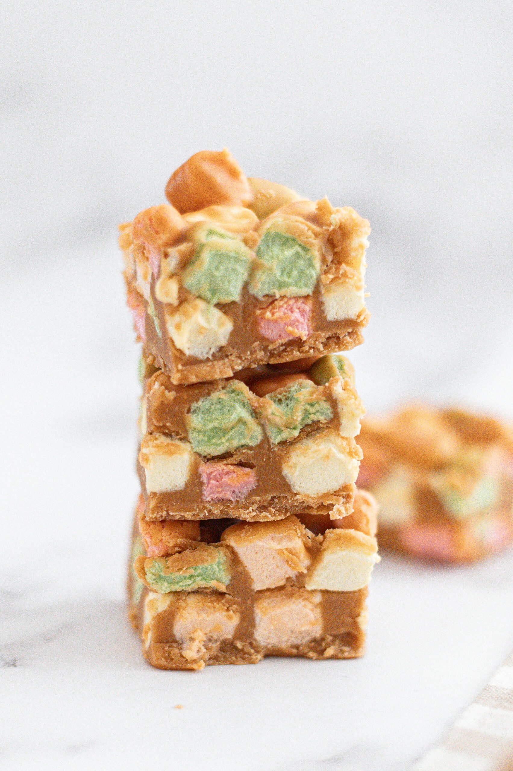Three squares of no-bake dessert bars with colorful mini marshmallows and a creamy tan base are stacked on a white surface, with a blurred bar in the background.
