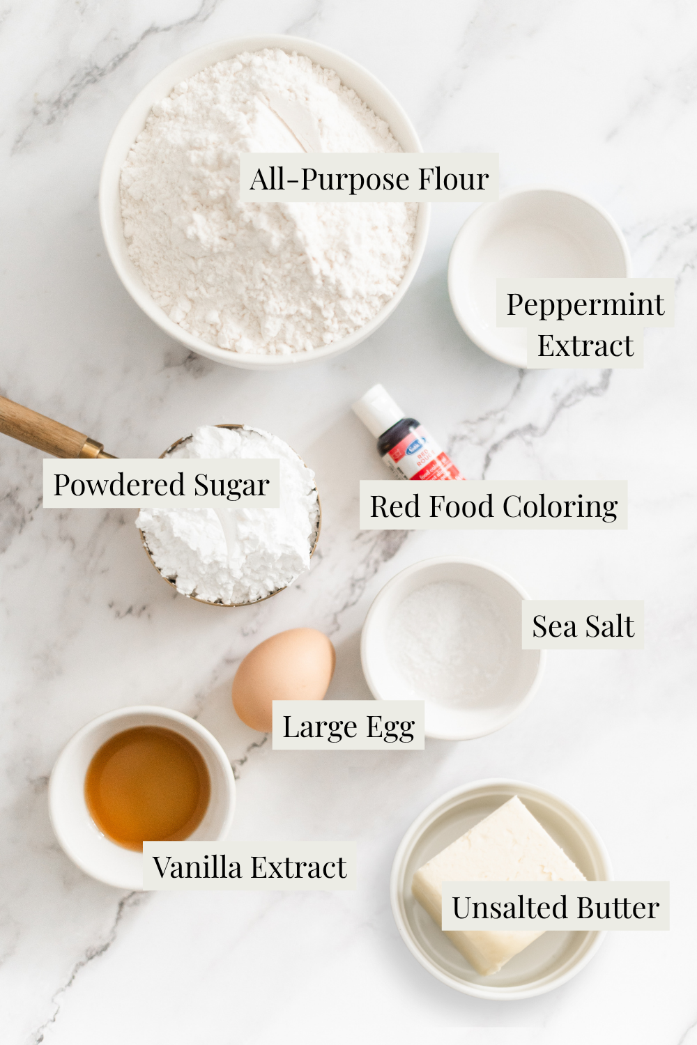 Flat lay of baking ingredients on a marble surface: all-purpose flour, powdered sugar, sea salt, unsalted butter, large egg, vanilla extract, peppermint extract, and a small bottle of red food coloring, each labeled.