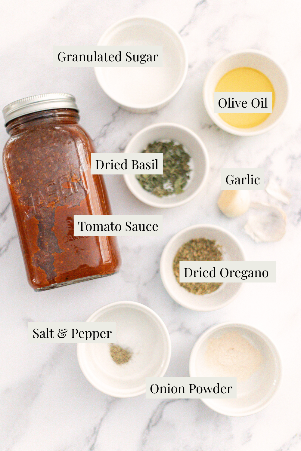 A jar of tomato sauce is surrounded by small bowls containing granulated sugar, olive oil, garlic, dried basil, dried oregano, salt and pepper, and onion powder, all labeled on a white marble surface.