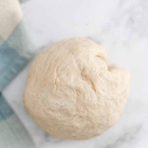 A ball of unbaked dough rests on a white marble surface next to a blue and beige striped cloth.