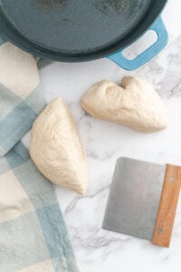Two portions of dough rest on a marble surface next to a dough scraper with a wooden handle, a blue checkered kitchen towel, and a blue-handled cast iron pot.