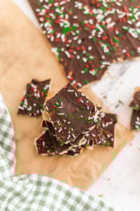 Squares of Homemade Christmas Crack (Saltine Toffee Bark) topped with red, white, and green sprinkles are stacked on parchment paper, with more pieces and a green checkered cloth nearby.