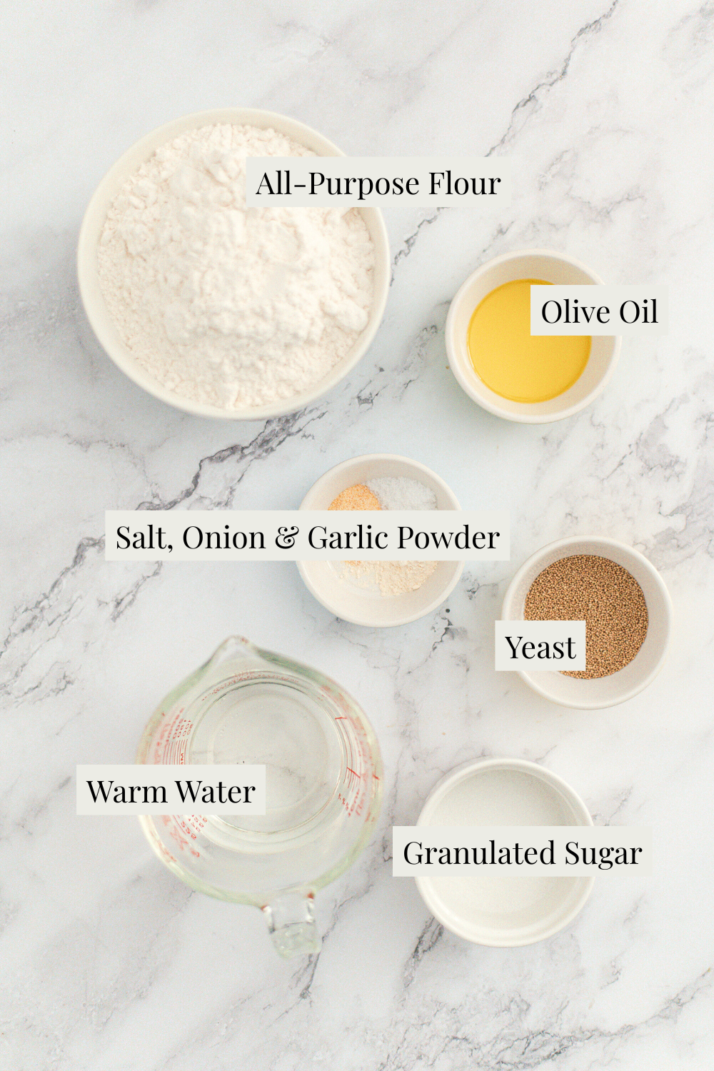 Five labeled ingredients are arranged on a marble surface: all-purpose flour, olive oil, salt/onion/garlic powder, yeast, warm water in a measuring cup, and granulated sugar in a small bowl.