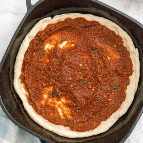 A cast iron skillet with pizza dough topped with red tomato sauce, ready to be baked. The skillet rests on a white marble surface next to a blue and white checkered cloth and a bowl of extra sauce.
