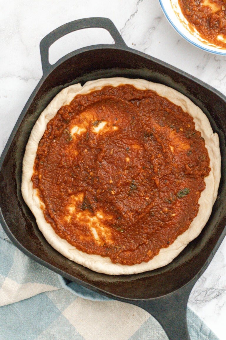 A cast iron skillet with pizza dough topped with red tomato sauce, ready to be baked. The skillet rests on a white marble surface next to a blue and white checkered cloth and a bowl of extra sauce.