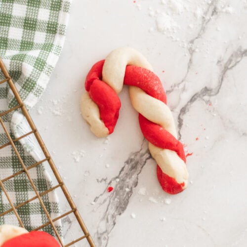 A red and white twisted candy cane-shaped cookie sits on a marble surface next to a green and white checkered cloth and part of a cooling rack.