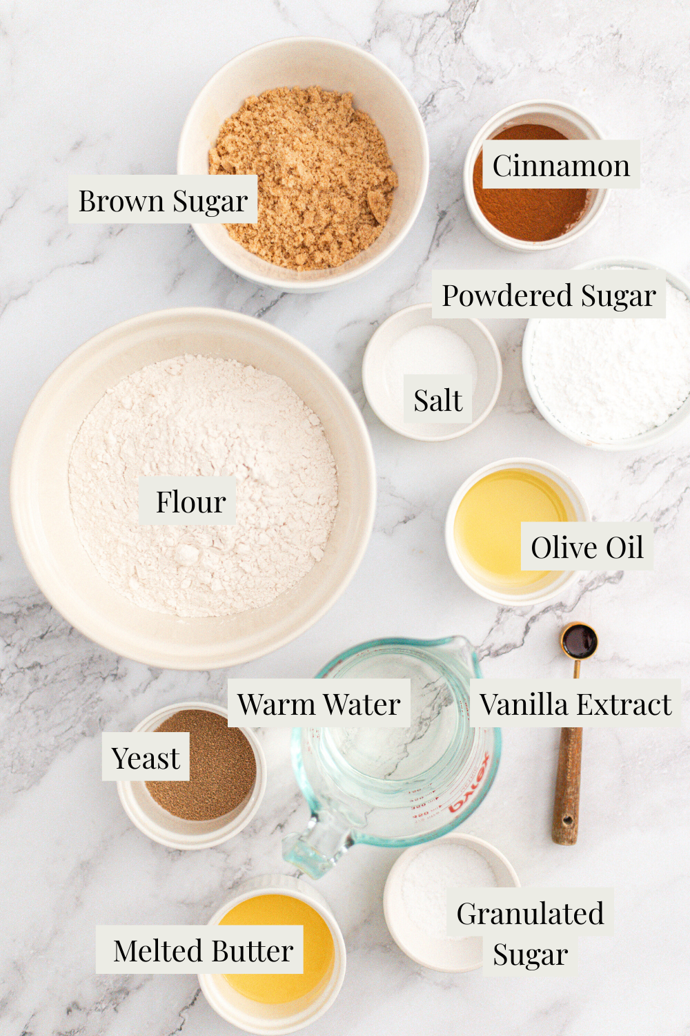 A marble surface with labeled bowls and cups containing brown sugar, cinnamon, powdered sugar, flour, salt, olive oil, vanilla extract, warm water, yeast, granulated sugar, and melted butter.