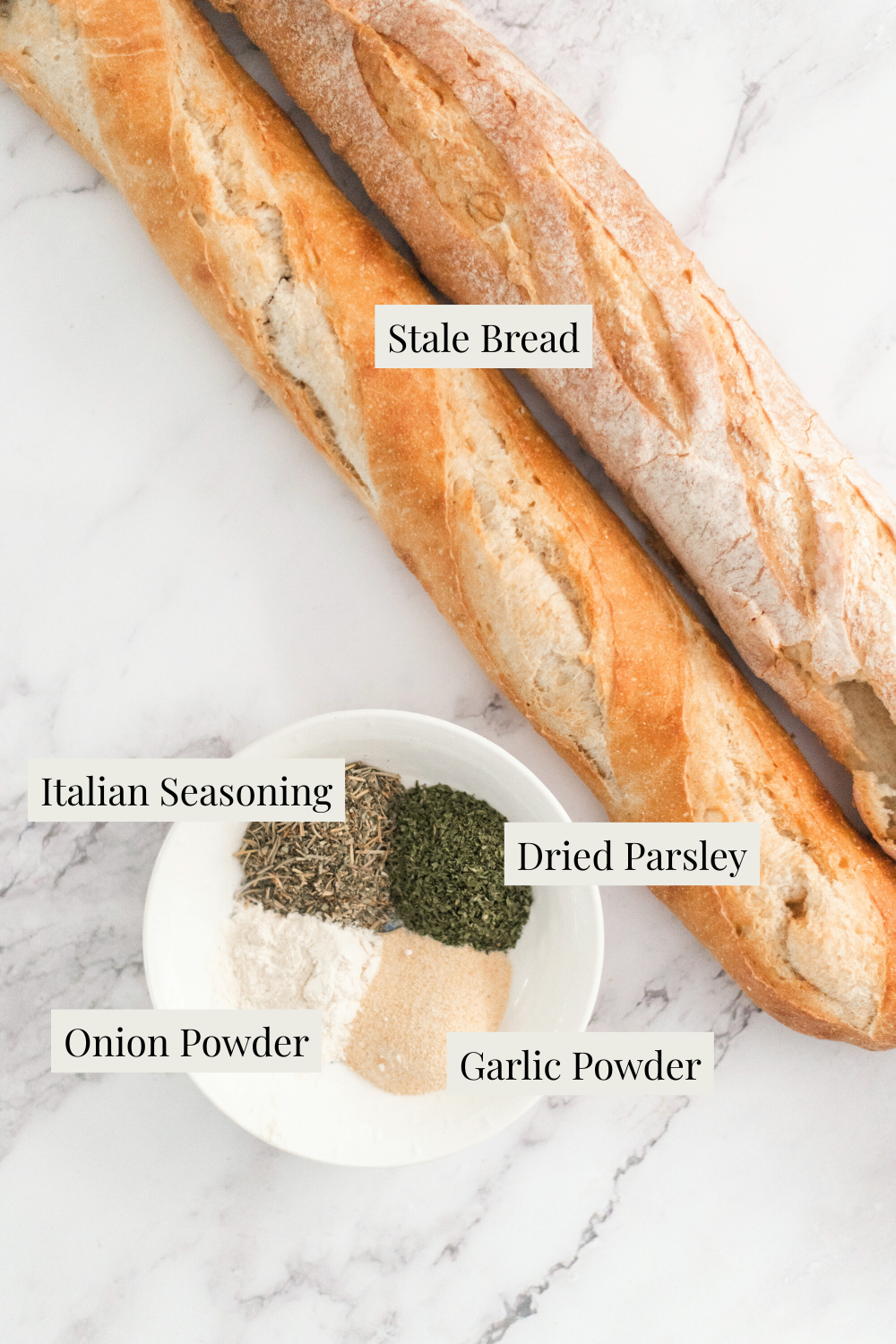Two baguettes labeled Stale Bread rest on a marble surface next to a bowl containing labeled piles of Italian seasoning, dried parsley, onion powder, and garlic powder—perfect ingredients for How to Make Homemade Breadcrumbs.