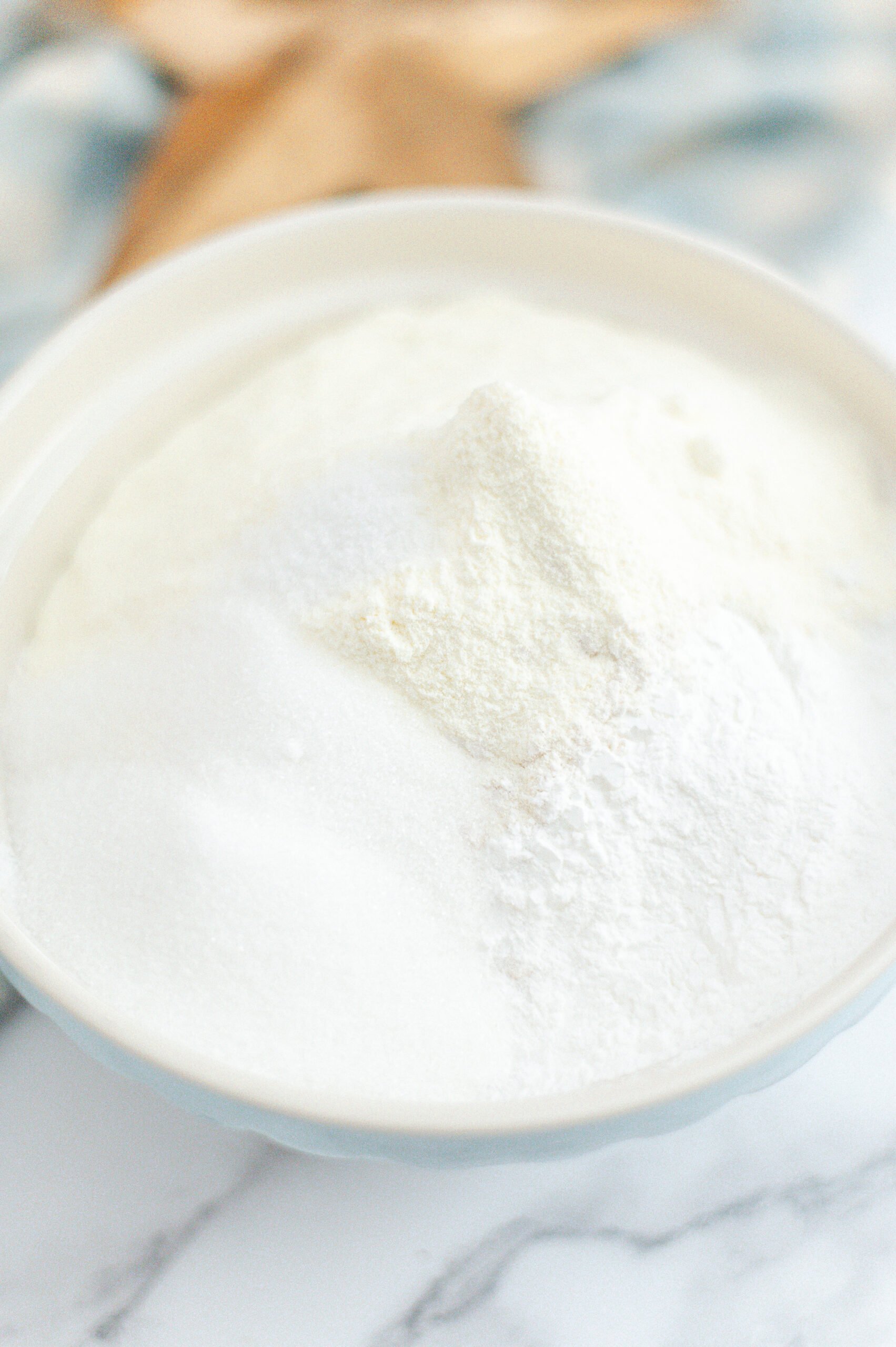 A close-up of a white bowl filled with a mound of white powders, likely sugar, flour, and possibly milk powder, on a marble surface. The background is softly blurred.