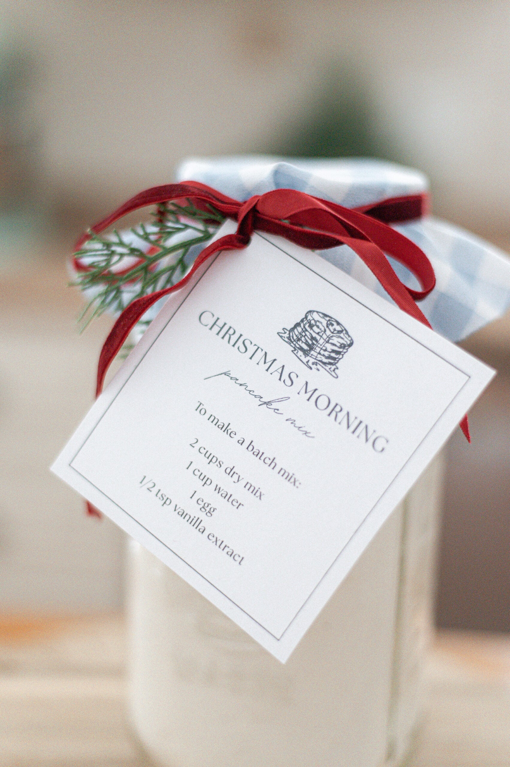 A glass jar with a blue-checkered cloth lid, tied with a red ribbon and a small pine sprig. A tag reads “CHRISTMAS MORNING pancake mix” with mixing instructions below.