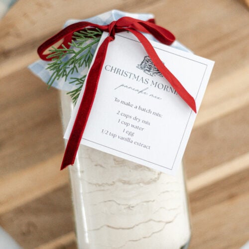 A jar of baking mix with a recipe card labeled Christmas Morning, tied with a red ribbon and small pine sprig, sits on a wooden board. The jar contains layered dry ingredients.