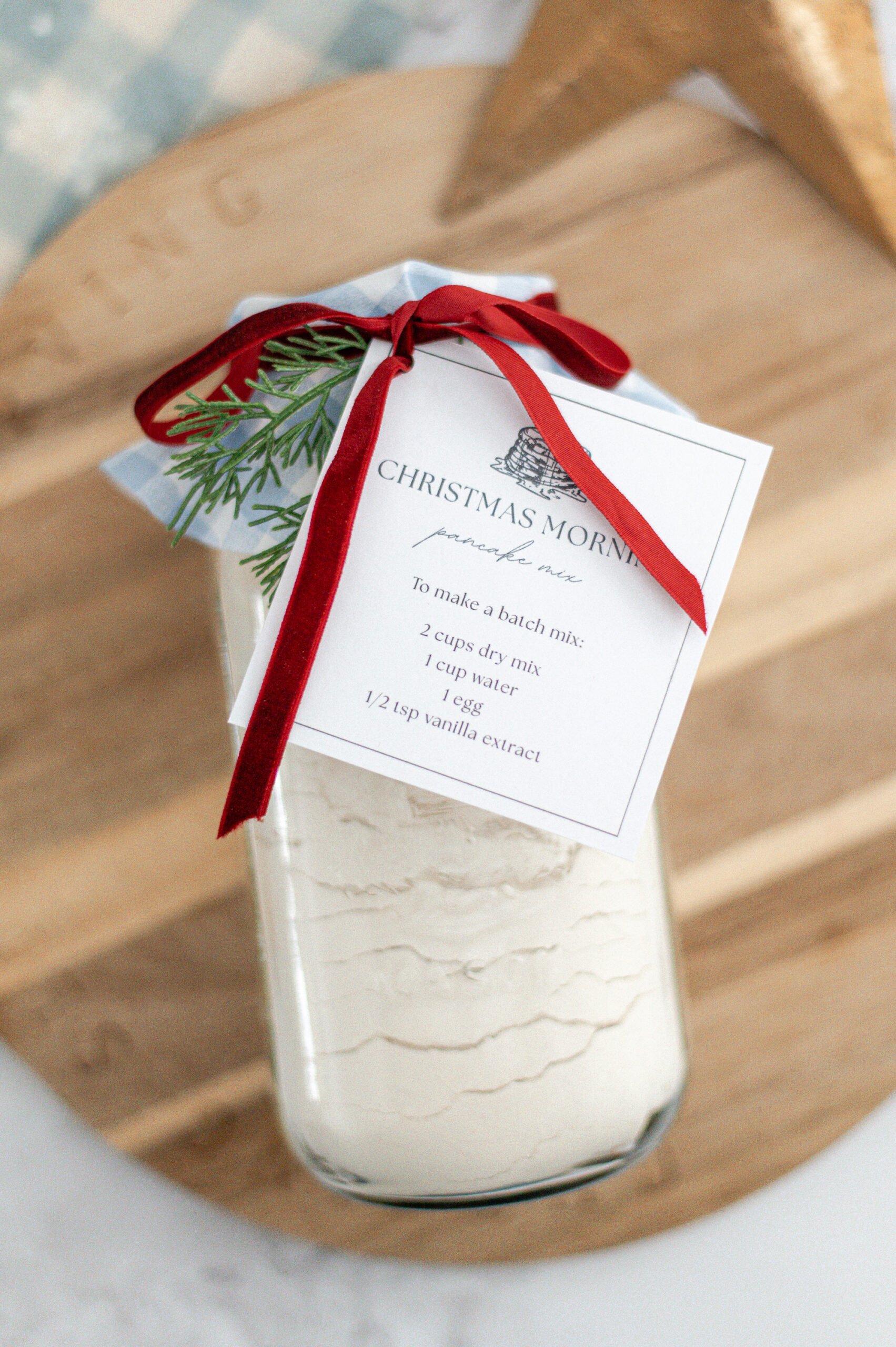 A jar of baking mix with a recipe card labeled Christmas Morning, tied with a red ribbon and small pine sprig, sits on a wooden board. The jar contains layered dry ingredients.