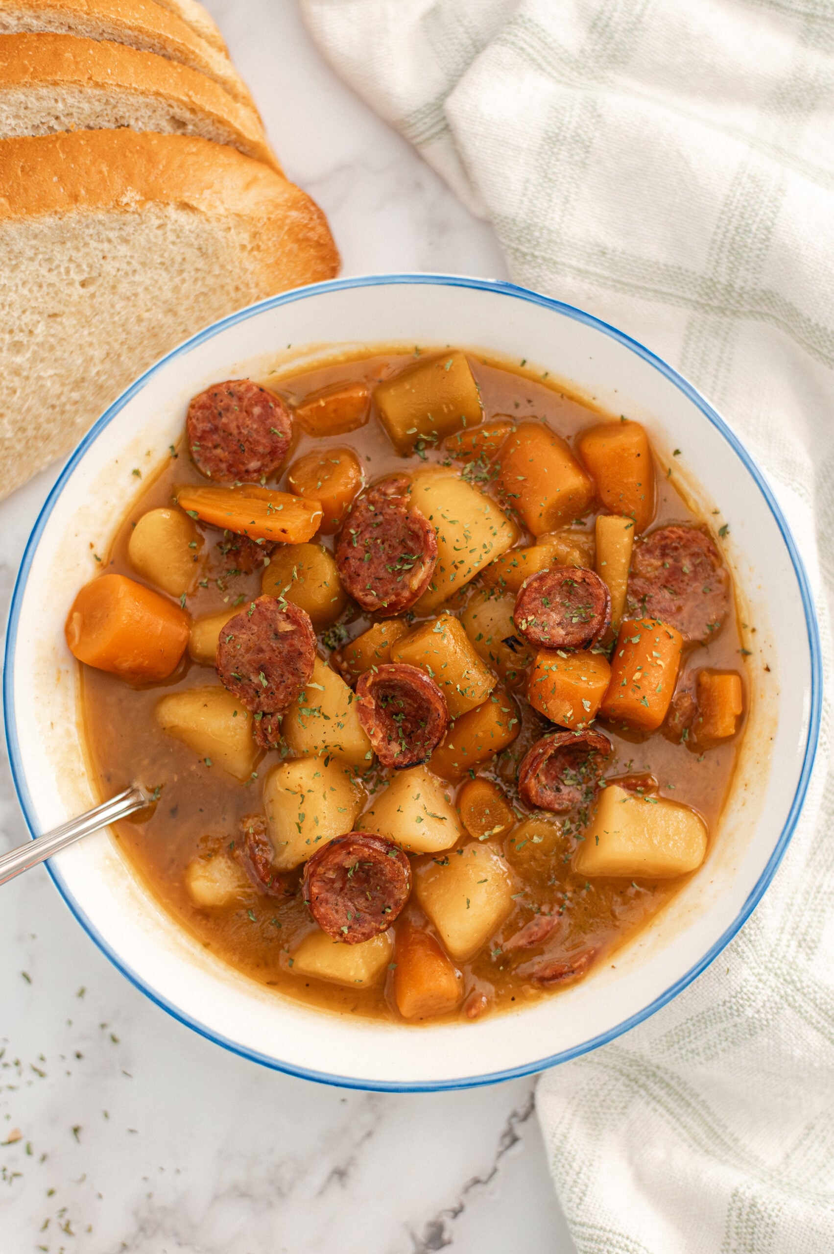 A bowl of hearty stew with sliced sausage, potatoes, and carrots in broth, garnished with herbs. A spoon rests in the bowl, with sliced bread and a white checkered cloth nearby on a marble surface.
